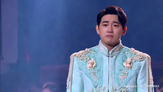 一个身穿浅蓝色挂满珍珠的华服的男歌手，听到感人的歌曲前奏后，回想到了之前出道时的重重困难，感慨万千蹲在舞台上用手遮住面孔掩盖抽泣，舞台下粉丝们很配合的大合唱 by Hailuo AI Video Generator