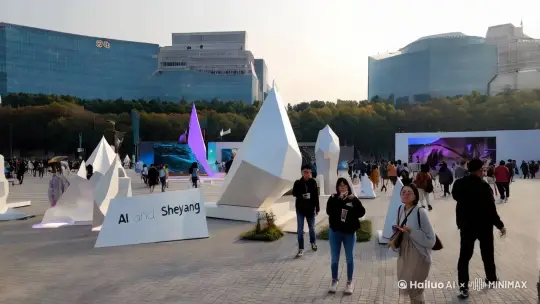 全景，射阳的文化广场上，一场以 “AI 与射阳” 为主题的创意展览正在举行。人们穿梭在各种 AI 创意作品之间，有利用 AI 技术制作的雕塑、摄影作品、音乐表演等，观众们纷纷拿出手机拍照留念，脸上充满了新奇和赞叹。 by Hailuo AI Video Generator