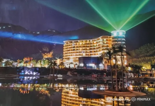 夜幕低垂，湖面如镜，一座座摩天大楼拔地而起，它们的轮廓被绿色激光勾勒，仿佛是夜空中最耀眼的星。远处的山峦被柔和的蓝色灯光渲染，宛如梦境中的幻境。湖边，一排挺拔的棕榈树轻轻摇曳，它们与近处的建筑一同在湖水中留下梦幻般的倒影。彩色灯光在背景中闪烁，营造出节日般的欢乐氛围。镜头缓缓移动，捕捉到楼上灯光忽明忽暗的变化，增添了几分神秘感。这是一幅充满现代都市魅力与自然美景交融的夜景画卷，中景至全景，平视角度，让人仿佛置身于这场光影盛宴之中。 by Hailuo AI Video Generator