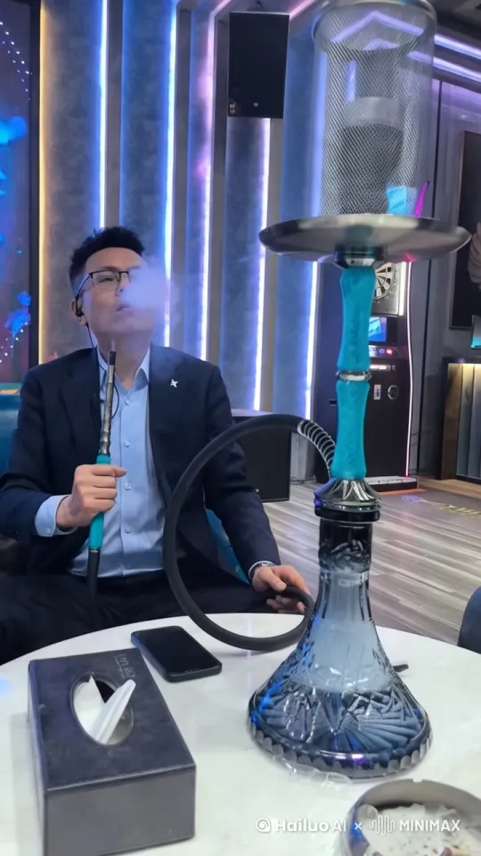 这张图片展示了一位正在使用水烟的人。水烟是一种传统的吸烟方式，通常在社交场合中使用。水烟壶的设计精美，常常成为室内装饰的一部分。从图片中可以看到，水烟壶的底座是蓝色的玻璃材质，上面有精美的花纹设计，整体看起来非常优雅。

此外，桌面上还有一个手机和一个纸巾盒，环境看起来像是一个现代化的室内场所，背景有彩色的灯光装饰，营造出一种轻松、愉悦的氛围。这种环境通常适合朋友聚会或休闲放松。

如果你有任何关于水烟或这种环境的具体问题，欢迎随时提问！ by Hailuo AI Video Generator