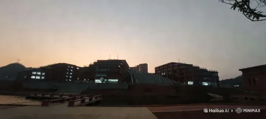 黄昏时分的校园风光视频，天空如同调色盘，渐变着橙蓝交响曲。教学楼灯火初上，倒映在静谧的水面，宛如梦幻泡影。镜头聚焦一条引领探索的石阶，两侧铁艺栏杆守护着学子们的步伐。远山剪影勾勒天际，一抹绿意从右侧闯入视野，树枝轻摇，似乎在诉说岁月的故事。随着太阳慵懒地探出头，一群群鸟儿振翅高飞，划破宁静的天幕，带来生机与希望。全景镜头，缓缓移动，捕捉每一刻光影变化，记录校园黄昏的温柔与活力。 by Hailuo AI Video Generator