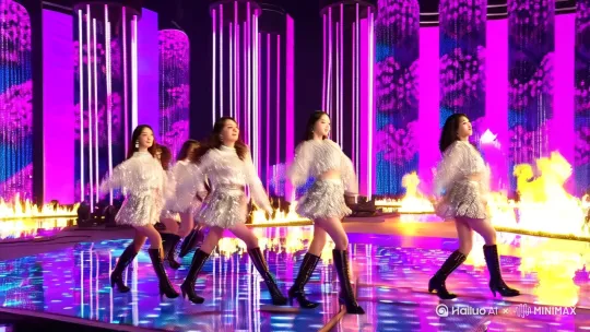 ：K-pop舞台中央，五名成员身穿银色亮片短裙与高筒靴，整齐排列的刀群舞动作，同步甩发与wave动作，舞台地板嵌入动态LED屏显示火焰特效，空中悬浮全息虚拟花瓣随节奏飘落，背景巨型环形霓虹灯管散射粉紫光晕，镜头特写成员妆容（猫眼眼线+渐变唇彩），8K超清质感，虚幻引擎渲染，电影级追光与慢镜头动态模糊 by Hailuo AI Video Generator