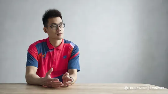 [推进,左移]一个帅哥在桌旁讲话，他慢慢看向镜头，扶了一下眼镜，眼睛充满智慧，露出微微的笑意，帅气，性感 by Hailuo AI Video Generator