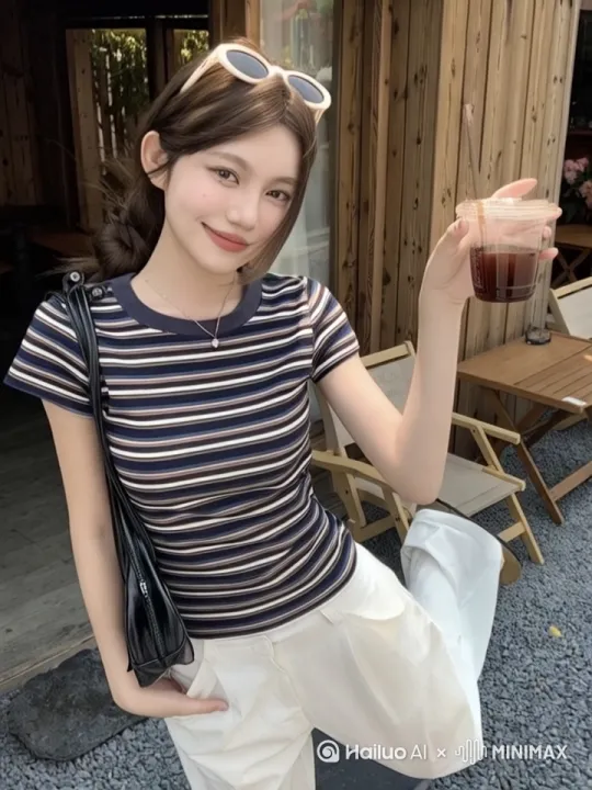 将图中时尚年轻女性制作成动态走路效果，她有棕色长发，用白色发卡别住一侧头发，戴着心形吊坠项链，身着图片中的衣服，背着浅绿色条纹大包，手里拿着透明塑料杯装的深色饮品。步伐自然，背景里木质建筑、桌椅等环境元素保留 by Hailuo AI Video Generator