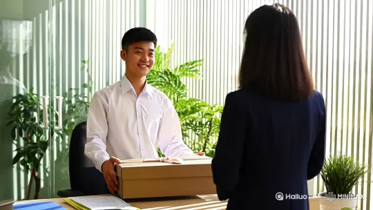 一位穿着白衬衫的青年教师站在教师办公室，面带微笑将礼品盒推回家长面前，透明玻璃隔断外可见绿植盆栽，阳光透过百叶窗形成光影条纹，办公桌上文件摆放整齐，画面采用固定镜头正面视角，自然光营造清新正直氛围。 by Hailuo AI Video Generator