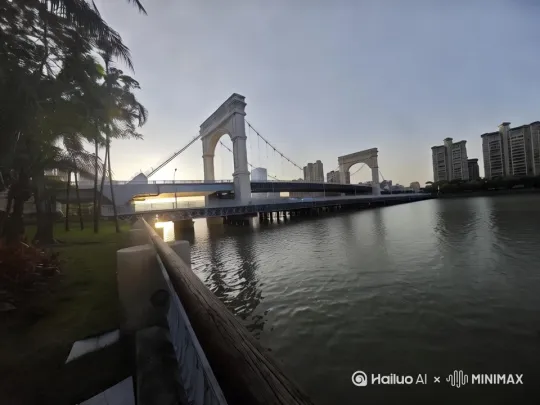 - 景别:全景
- 画面:从稍远位置,完整呈现桥梁横跨河流的全貌,夕阳在画面一侧,将天空染成浅橙与淡紫渐变,河水波光粼粼,岸边树木、草地和远处建筑作为背景层次分明。
- 运镜:固定镜头,缓慢拉开一点点(模拟人站定后视野自然展开),让画面从初始的聚焦桥梁,逐渐纳入更多周边环境。
- 声音:轻柔的风声,河水流动的潺潺声,远处若有若无的城市嘈杂声(后期处理得很淡,作为环境底噪)
- 拍摄建议:选在黄昏日落前20分钟左右,光线柔和时拍摄。使用广角镜头,三脚架固定,保证画面稳定。 by Hailuo AI Video Generator