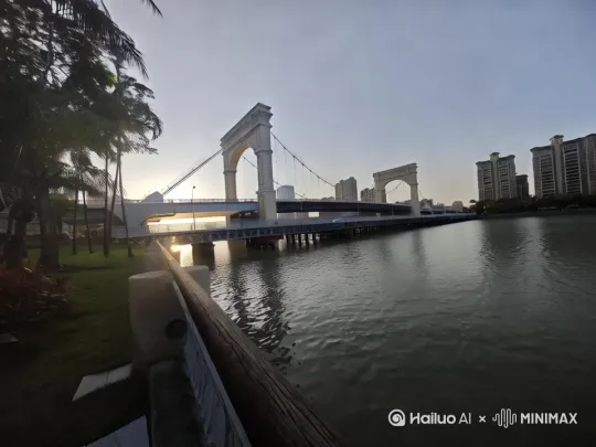 - 景别：中特写
​
- 画面：聚焦桥梁的主体结构，比如那两座白色拱形门，展现建筑细节，阳光在桥身投下的阴影，以及连接的钢索线条。河水在桥下形成倒影，随着水波微微晃动。
​
- 运镜：缓慢推镜头，从桥梁全景向拱形门推进，突出建筑质感，过程中轻微晃动镜头（手持稳定器，模拟人轻轻向前走两步观察的视角，带点呼吸感）。
​
- 声音：风声稍大一点，能听到树叶被吹动的“沙沙” 声，河水拍击桥墩的细微声响
​
- 拍摄建议：调整角度，让阳光侧照桥梁，凸显立体感。可后期稍微加强桥身的明暗对比，突出细节。 by Hailuo AI Video Generator