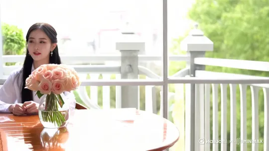 一片明亮的阳台，镜头从桌面镜头开始慢慢推进，拍摄到木桌前明亮的环境里坐着一个人，在这个人左手边桌子上有一束淡粉色玫瑰花，女人嘴在说话，女人留着黑色的长发，穿着白色的衬衣 by Hailuo AI Video Generator